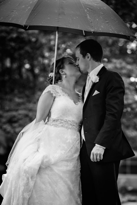 Black and white wedding photo of a bride and groom walking in the rain under an umbrella. Photo taken on the campus of Augsburg College in Minneapolis, MN.