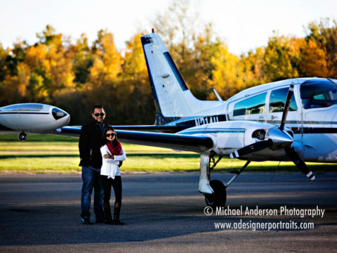 A cute couple posing by a small airplane at the Anoka County Airport during their fall color engagement portraits.