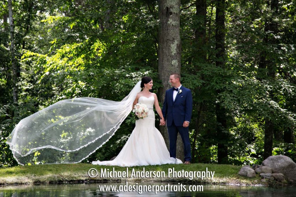 A very pretty wedding photograph of the bride and groom with her veil flowing in the wind; Image taken by a pretty pond before their Grand View Lodge destination wedding.
