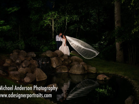 Stunning nighttime wedding photo of the groom and his bride with her veil flowing in the breeze. Wedding photo taken at a waterfall and pond at Grand View Lodge in Nisswa, MN.