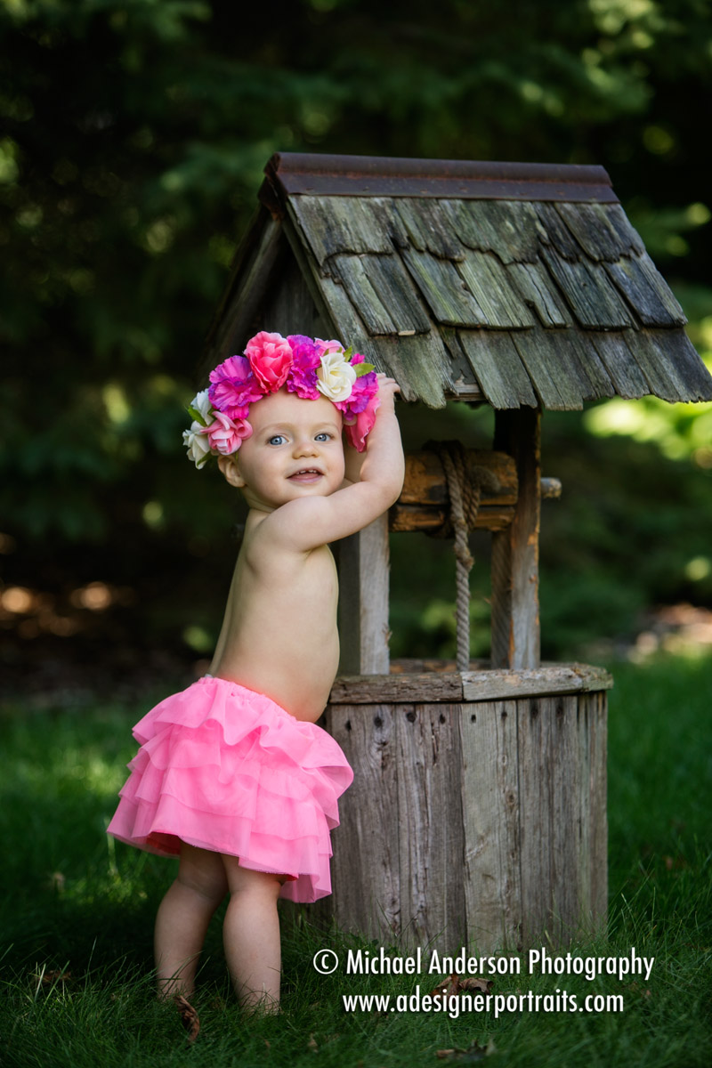 Cute one year old baby girl portraits of an adorable girl dressed in a pink skirt and a flower headband. She's standing by a small wishing well at Anderson's Portrait Park in Mounds View, MN.