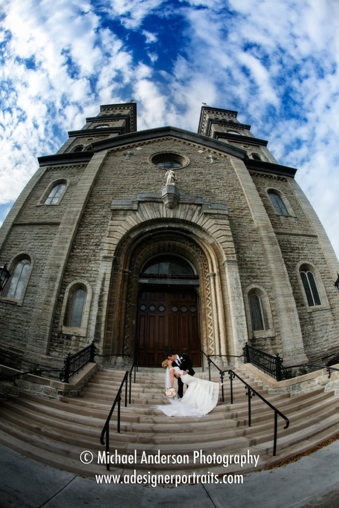 The bride and groom kissing while the groom is dipping her. Beautiful wedding photograph taken with a fisheye lens on the front steps at Church of the Assumption in St. Paul, MN.