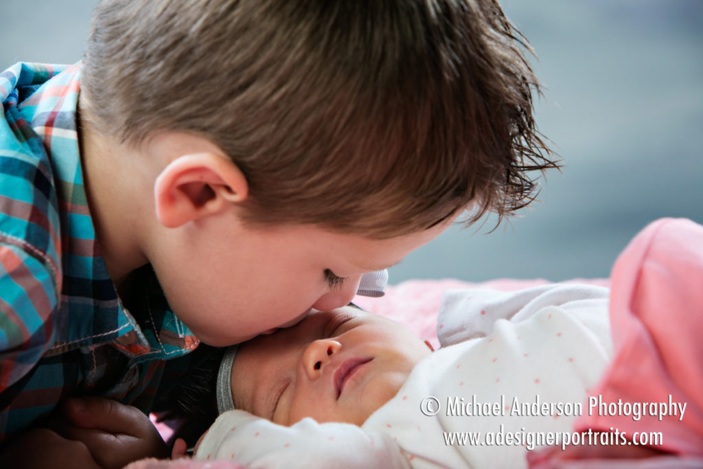 A photo studio portrait of a big brother kissing his new sister, an adorable newborn baby girl.