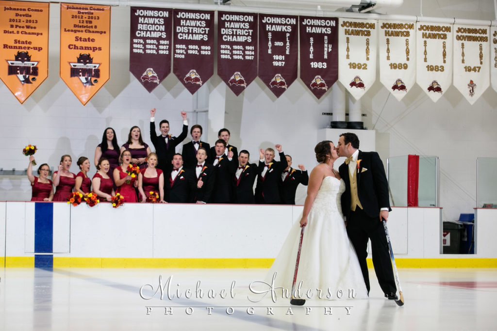The wedding party celebrates from behind the hockey boards watching the bride & groom share a kiss on the ice. A very unique wedding photo taken at Saint Paul Johnson High School on the East Side of St. Paul, MN.