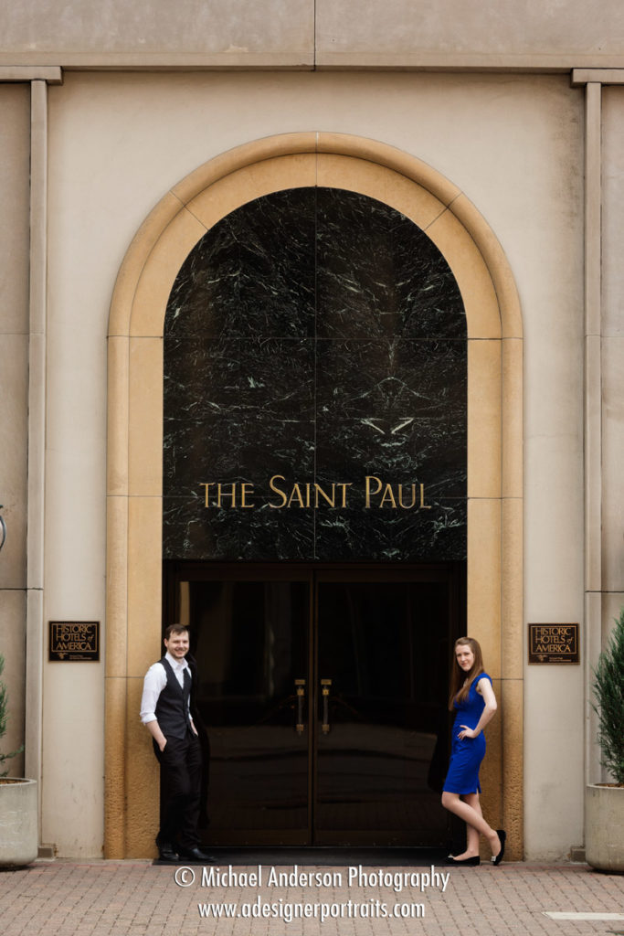 A cute couple posing by the doors of the hotel's historic street level entrance. Image creating during their engagement portraits at The Saint Paul Hotel.