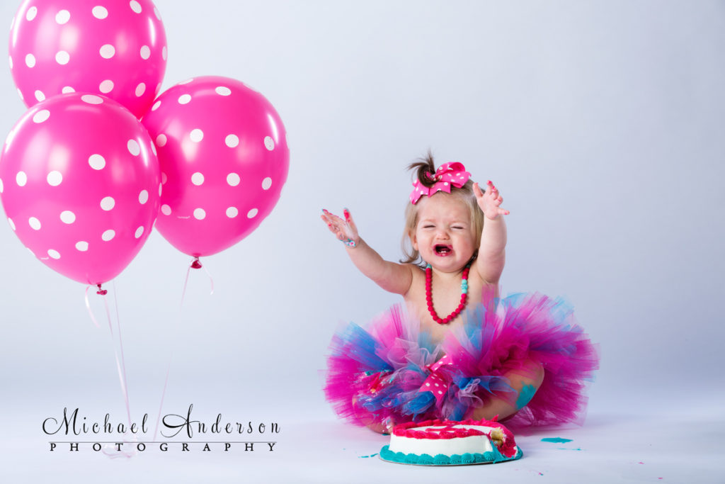 One-year portraits of Lainey, crying by her birthday cake and pink balloons.