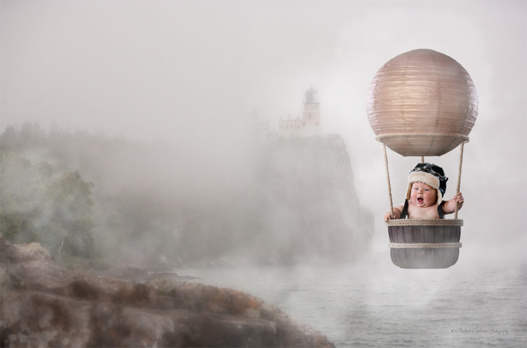 A cute fantasy baby portrait titled "The Little Aviator." An adorable five month old baby boy flying a hot air balloon in the fog over Lake Superior with the famous Split Rock Lighthouse in the background.