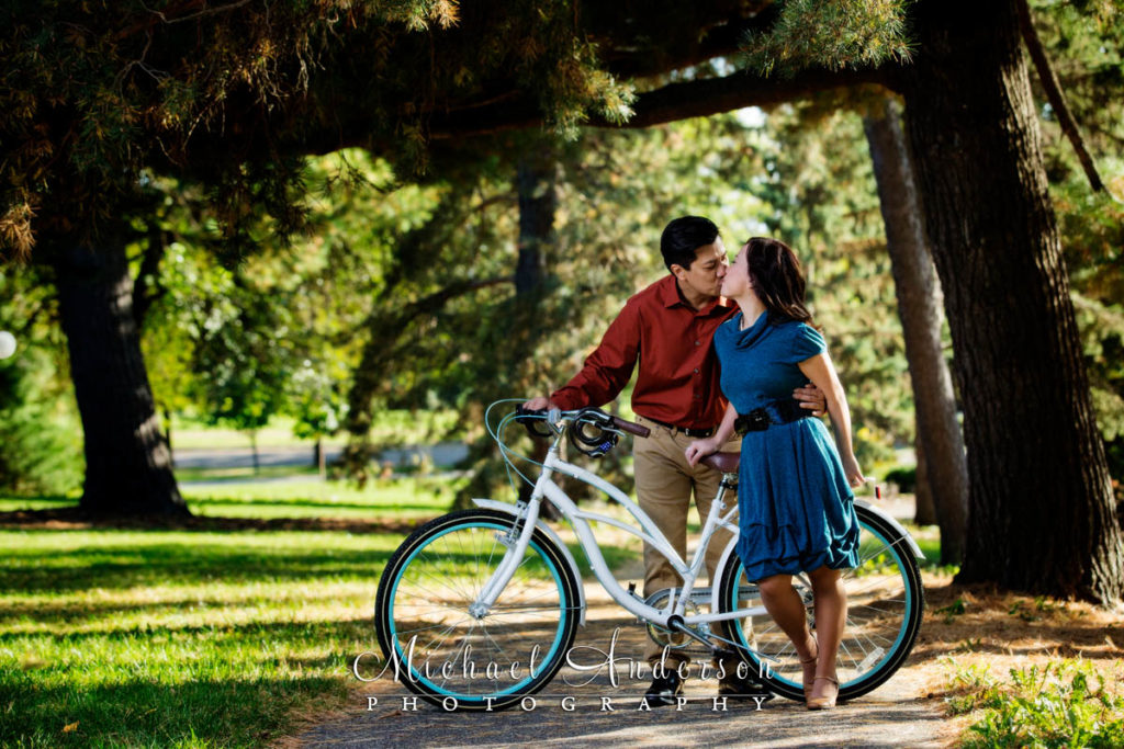A cute couple sharing a kiss on a white bicycle, in a very pretty setting at Como Park. The very cute image was taken during their Como Park engagement portraits.