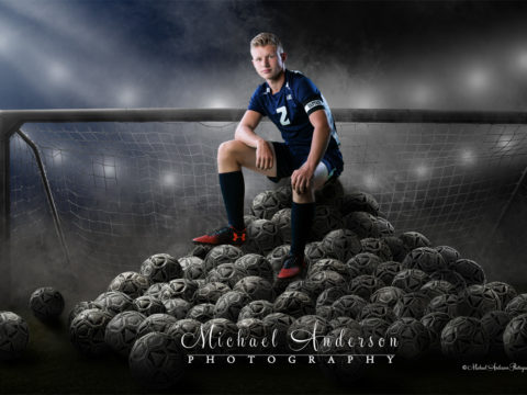 Mounds View professional photographer. Senior portrait on a big pile of soccer balls. Josh is sitting in front of a soccer goal and the composite takes place at night in a stadium with lots of smoke.