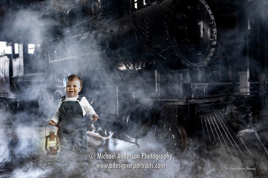 Baby Boy Train Engineer Photos. A super cute one year old baby boy holding a train lantern and dressed as a train engineer. A green screen composite photo with a steam locomotive at train station with dramatic lighting and steam.