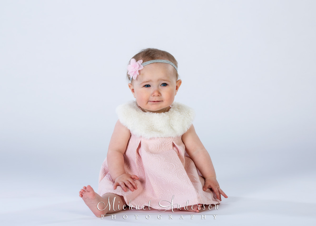 A simply adorable studio portrait of a nine-month-old baby girl wearing a cute pink dress.