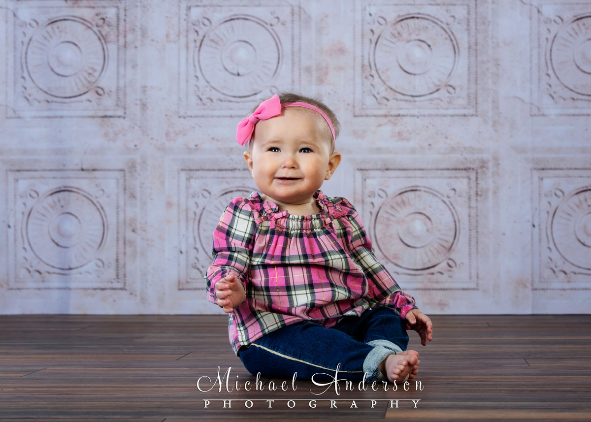 A simply adorable studio portrait of a nine-month-old baby girl wearing a pink plaid outfit.