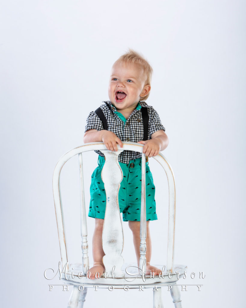 A cute one-year-old boy standing a white chair.