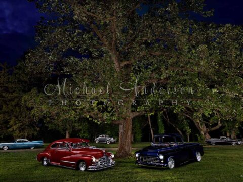 A stunning light painting of a 1939 Packard Coupe 110 Series, a 1948 Pontiac Silver Streak, a 1955 Chevrolet 3100 Stepside Shortbox pickup truck, a 1959 Cadillac Coupe Deville, and a 1959 Chevy El Camino under a huge oak tree at the client's home.