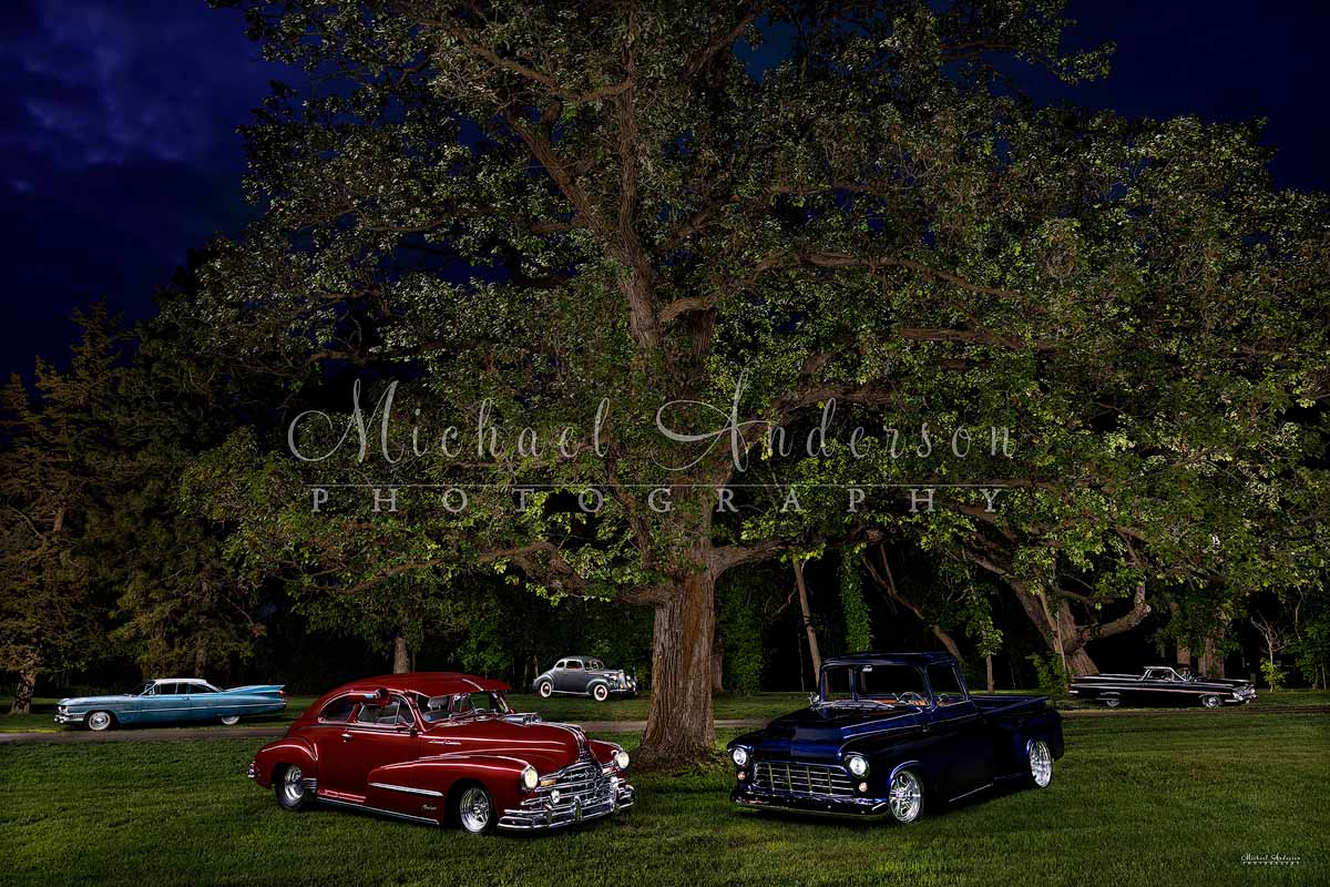 A stunning light painting of a 1939 Packard Coupe 110 Series, a 1948 Pontiac Silver Streak, a 1955 Chevrolet 3100 Stepside Shortbox pickup truck, a 1959 Cadillac Coupe Deville, and a 1959 Chevy El Camino under a huge oak tree at the client's home.